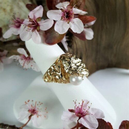 Elegant gold ring with a floral design on a white stand surrounded by delicate pink blossoms.